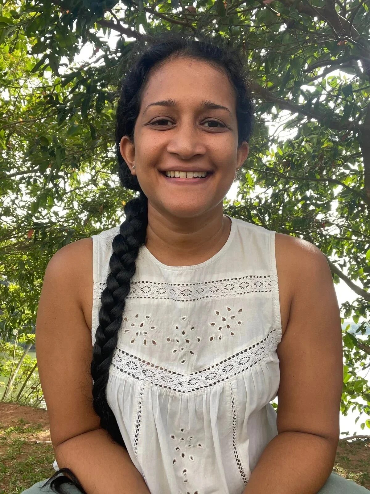 A woman smiling outdoors with green trees in the background, wearing a white sleeveless top with embroidered holes and a long dark braid.