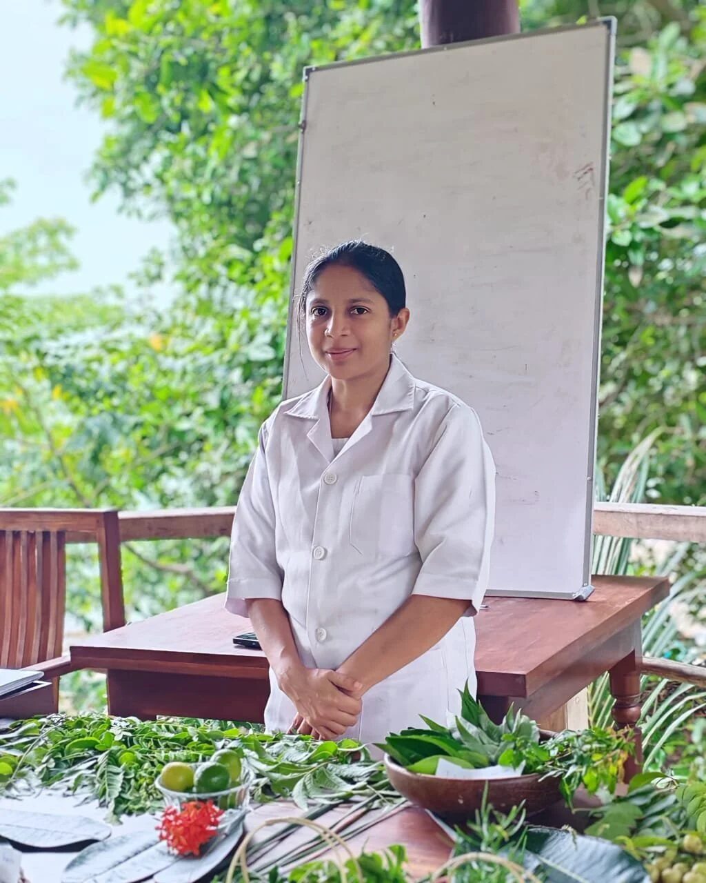 Young girl in a white coat standing behind a wooden table outdoors, with leafy green trees in the background, on a porch or patio area.