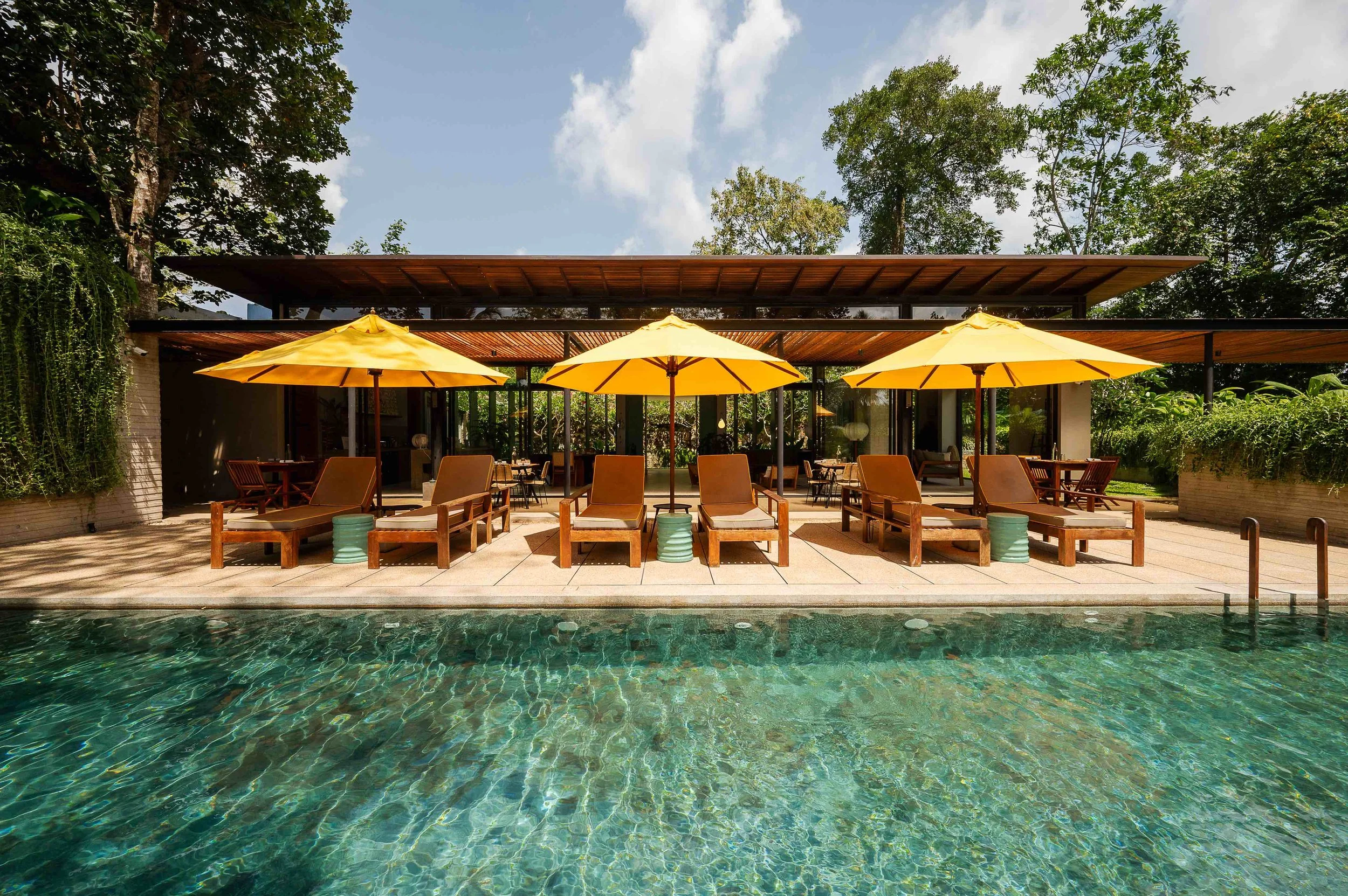 Poolside patio with yellow umbrellas, wooden chairs, and lush green trees in the background.