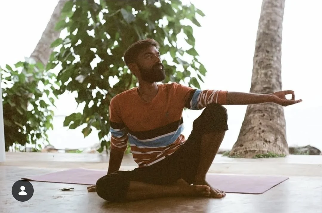 A man with a beard and short hair doing yoga on a mat outdoors near trees, sitting cross-legged with one arm extended forward and the other supporting him.